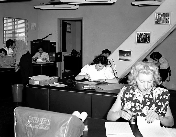 Black-and-white photograph of an office from the mid-20th century. Several women and men are working at desks in a shared workspace. In the foreground, a woman with light-colored hair sits at a desk reading or organizing papers. Behind her, other women are seated at desks, writing or handling documents. On the left side of the room, a man leans over a desk speaking to a seated woman, while another man works at a desk in the background. The room contains filing cabinets, papers, and office equipment. Framed photographs hang on the wall near a staircase, and the overall atmosphere suggests a busy administrative or clerical office.